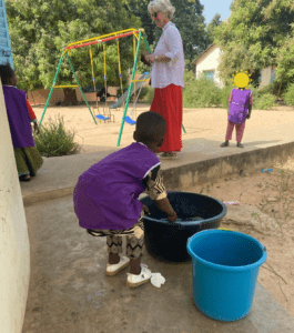 Au premier plan un enfant de dos attrape une bassine d'eau, il ou elle porte une blouse d'école. Au deuxième plan Marie-Hélène Aubert, Vice-Présidente d'YCID discute avec un enfant.